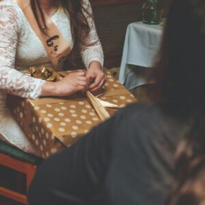 A woman at a bridal shower unwrapping a polka dot gift indoors, wearing a sash.