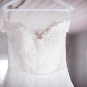 Beautiful lace wedding dress on a hanger in a softly lit room, showcasing intricate design.