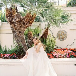Bride elegantly seated in garden with flowers and palm tree, wearing a luxurious white wedding dress.